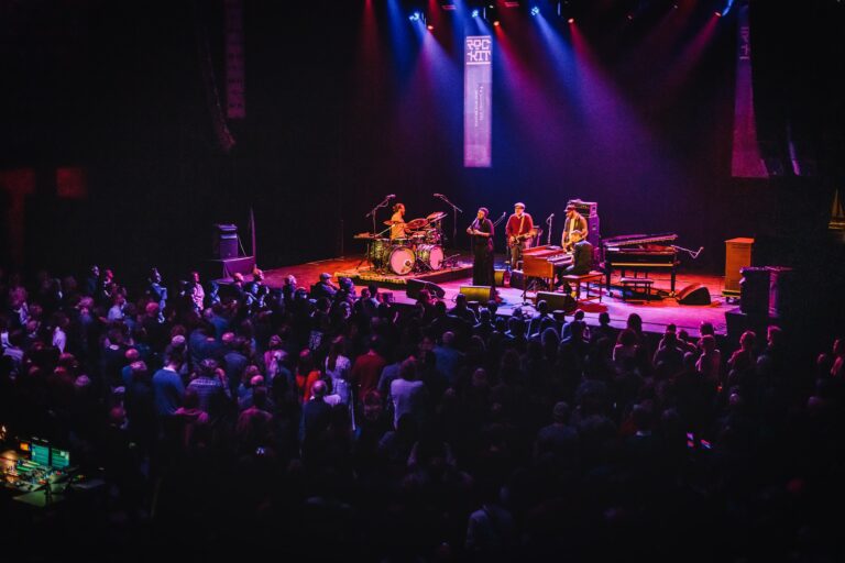 Een band speelt op een podium voor een enthousiast publiek, verlicht door kleurrijke lichten.