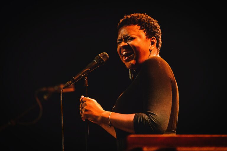 Een vrouw met kort haar zingt emotioneel in een microfoon, met warme verlichting op het podium.