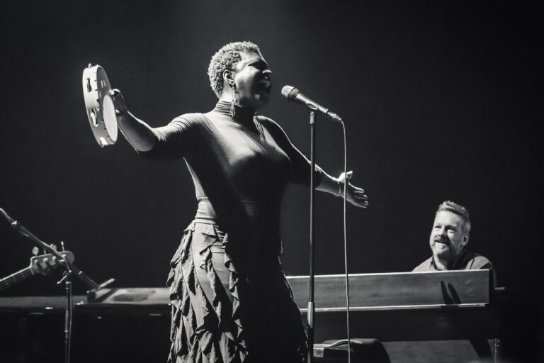 Zangeres met tamboerijn in de hand tijdens een optreden, met een pianist op de achtergrond.