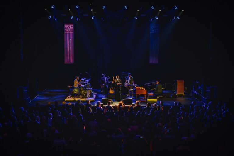 Een band treden op op een podium voor een enthousiast publiek, verlicht door een kleurrijke lichten.