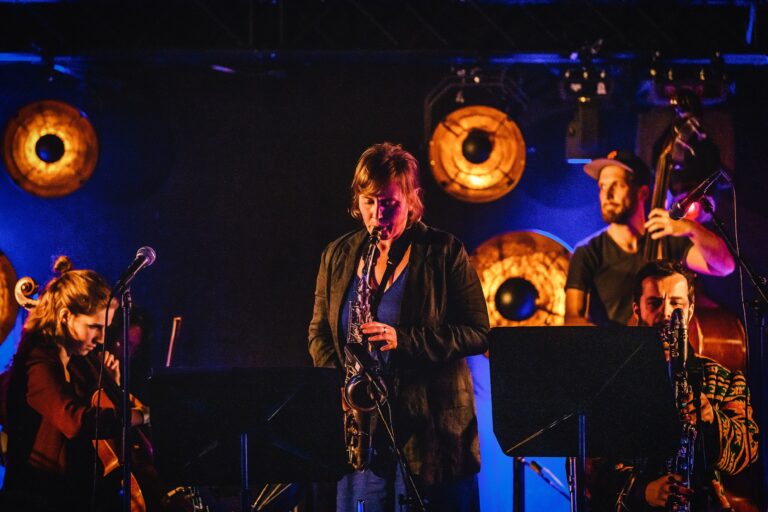 Een muzikante zingt op een podium met muzikanten op de achtergrond, omringd door sfeerlicht.