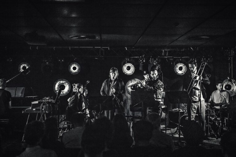 Een live muziekoptreden met artiesten op het podium, verlicht door ronde lichten in een donkere setting.