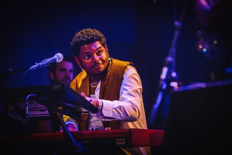 Een muzikant speelt op een keyboard tijdens een optreden, omringd door een sfeervolle, met licht verlichte achtergrond.