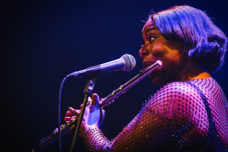 Een muzikante speelt een fluit terwijl ze in een microfoon zingt, verlicht door sfeervolle lichten.