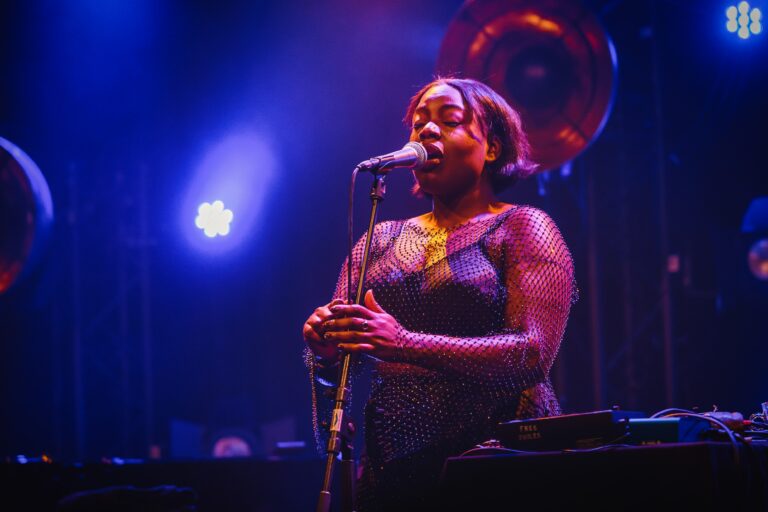 Zangeres staat op het podium met een microfoon, omringd door kleurrijke lichten.