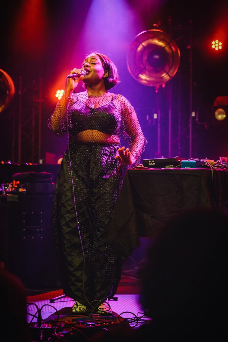 Een singer-songwriter staat op een podium en zingt, omringd door kleurrijke verlichting.