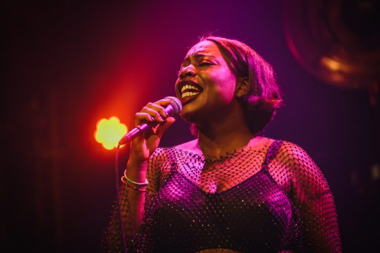 Een vrouw zingt enthousiast in een stijlvolle outfit tegen een achtergrond van gekleurde lichten.