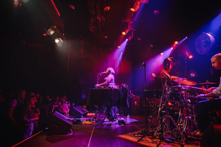Een live muziekoptreden met een artiest achter een synthesizer en een drummer, omgeven door sfeervolle verlichting en een enthousiast publiek.
