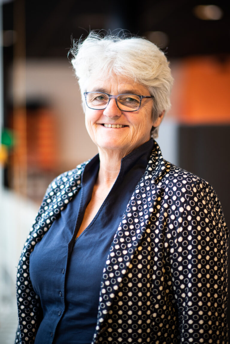 Een vrouw met grijs haar en een bril, gekleed in een gepatternde blazer, glimlacht naar de camera in een zakelijke setting.