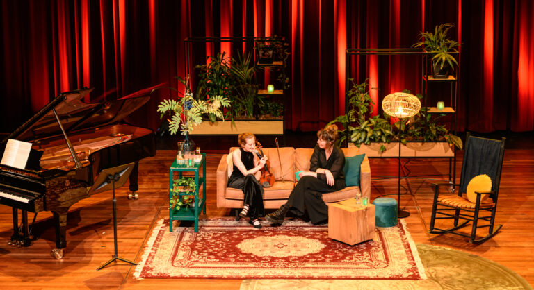 Twee vrouwen zitten op een bank in een sfeervolle, plantenrijke setting met een piano op de achtergrond.