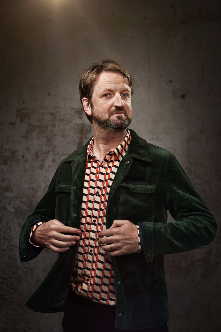 Een man met een baard draagt een groene jas en een kleurrijk gestreept shirt, terwijl hij naar de camera kijkt.