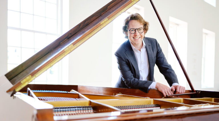 Pianist Hannes Minnaar die lachend op een houten vleugel leunt.