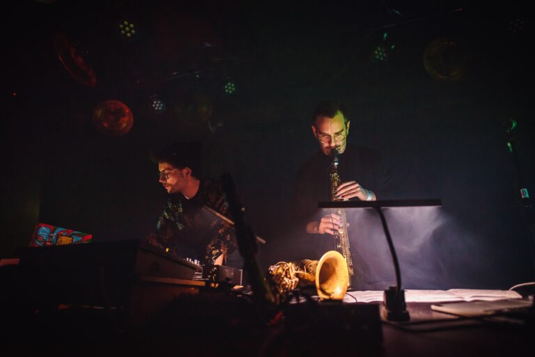 Twee muzikanten voeren een live optreden uit met een saxofonist en een toetsenist in een sfeervolle, gedimde omgeving.