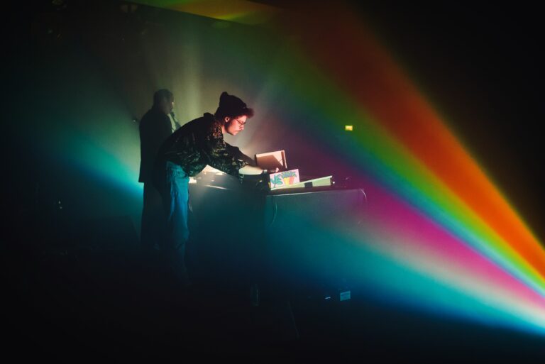 Een muzikant staat achter een tafel terwijl kleurrijke lichtstralen de ruimte verlichten.