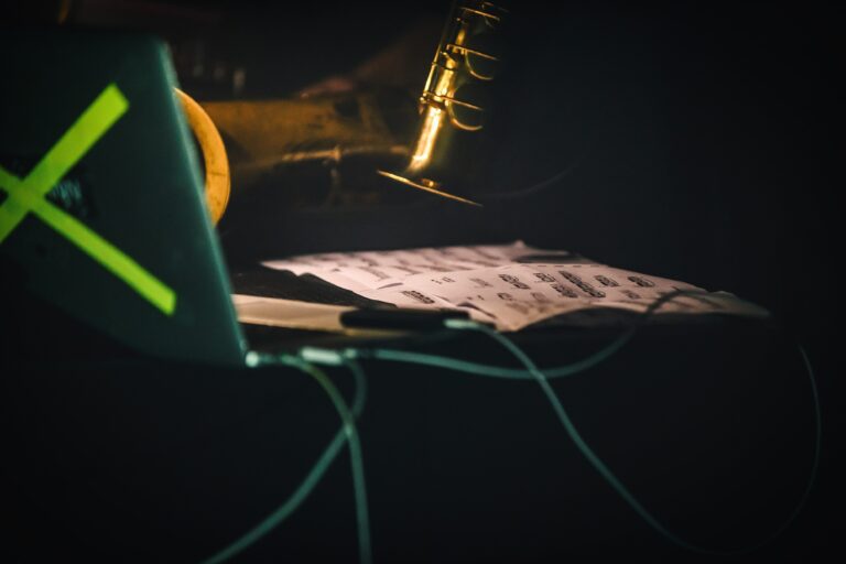 Een laptop naast bladmuziek met een microfoon op de achtergrond in een donkere omgeving.