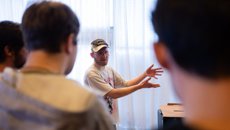 Een man met een pet en bril geeft een uitleg aan een groep mensen in een lichte ruimte met gordijnen. Hij gebaart met zijn handen terwijl de groep hem aandachtig bekijkt.