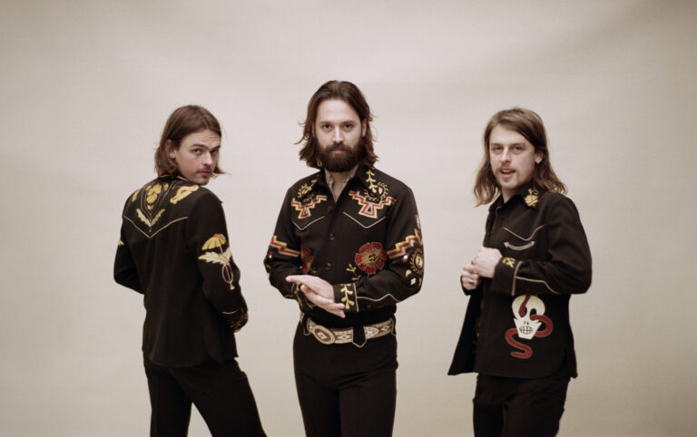 De drie bandladen van DeWolff, die volledig in het zwart gekleed voor een witte achtergrond staan.