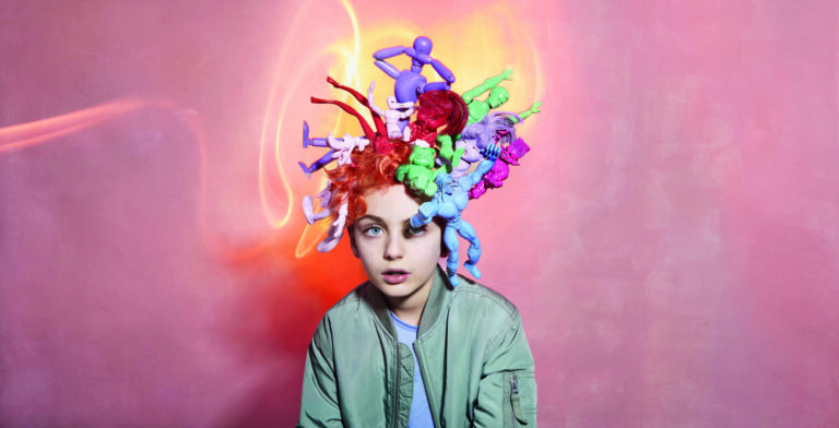 Roze achtergrond met jongentje met groen jasje met allemaal speelgoedpoppen uit zijn hoofd