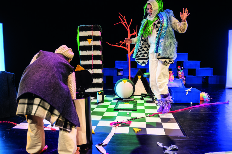 Een kleurrijke theaterscene met acteurs in opvallende kostuums op een speels, gestileerd podium.