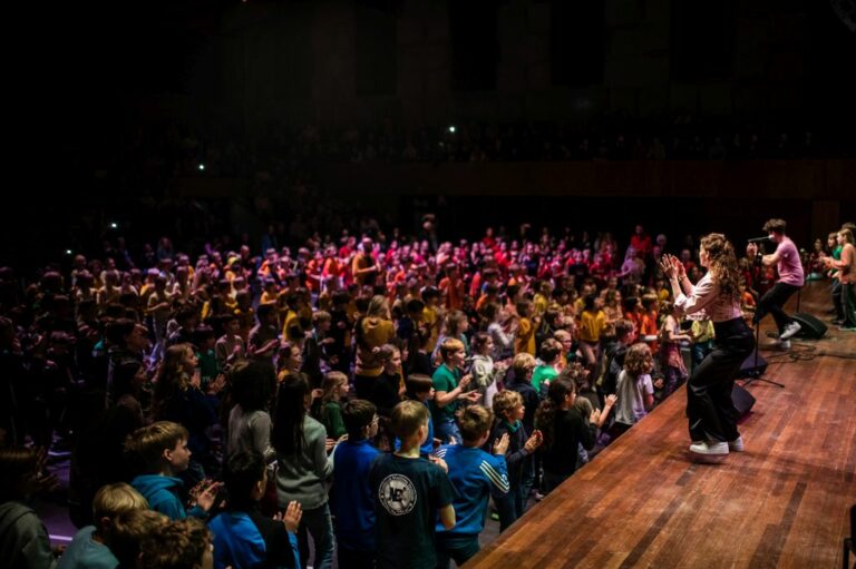 Een groot publiek applaudisseert terwijl een spreker op het podium staat, omgeven door kleurrijke lichten.
