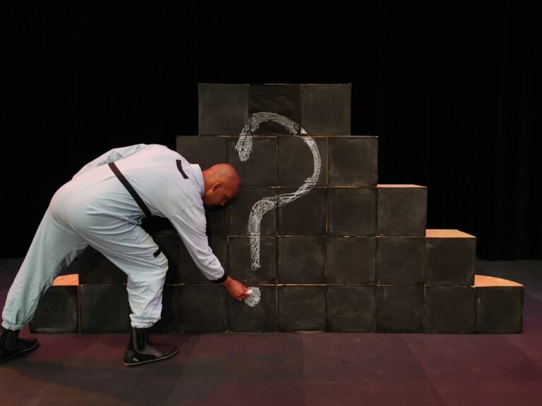 Een man in een grijs kostuum tekent een vraagteken op een muur van zwarte blokken.