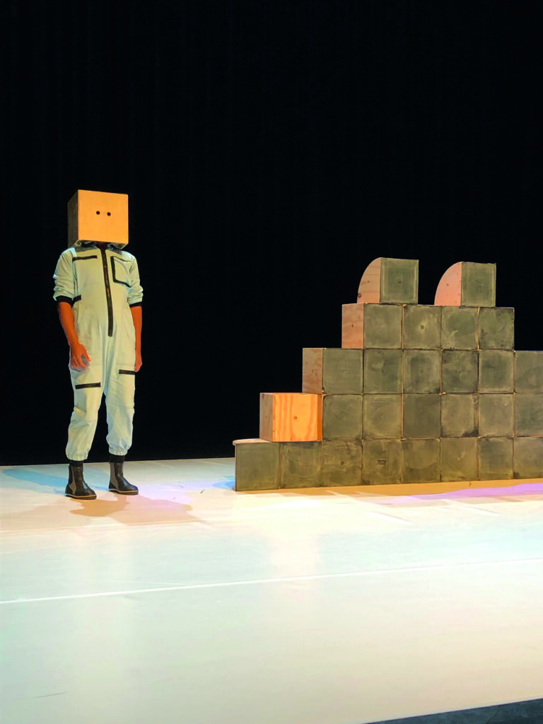 Acteur met doos op hoofd naast een stapel dozen