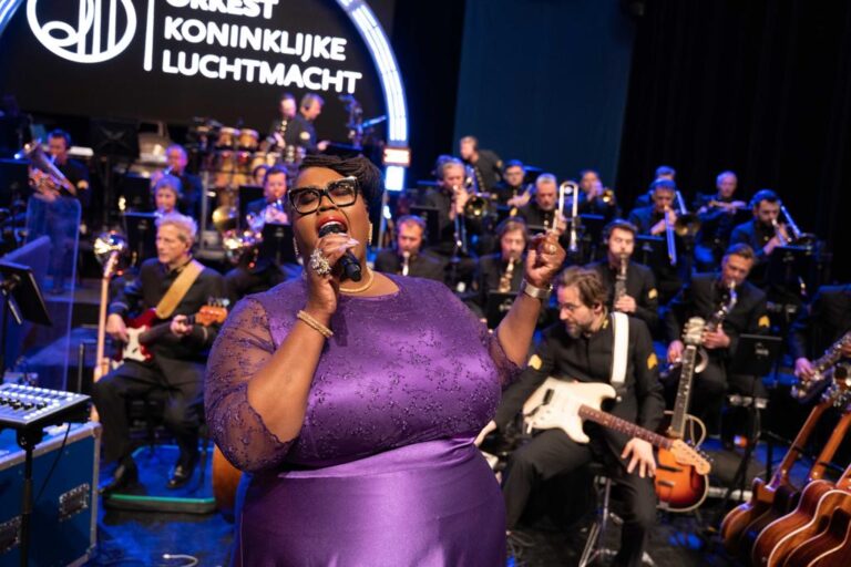 Een zangeres in een paarse outfit staat op het podium voor een groot orkest, terwijl ze de microfoon vasthoudt en zingt.