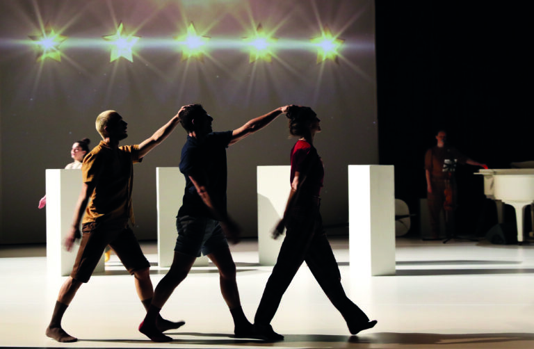 Een groep dansers beweegt zich over het podium onder heldere lichten, met enkele witte platforms op de achtergrond.