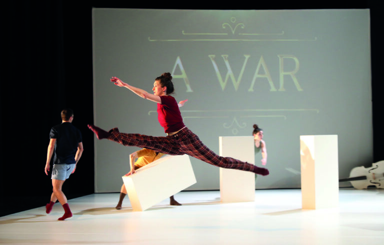 Een danseres maakt een sprongetje over enkele obstakels op het podium, met een achtergrond die de tekst 