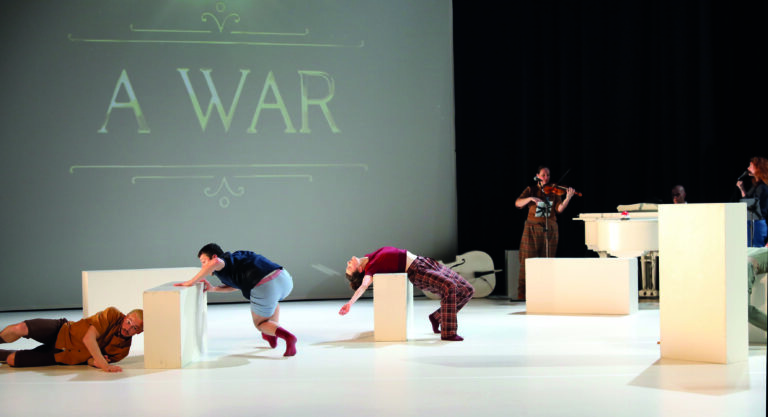 Een podium met dansers en muzikanten, waarbij de dansers zich over blokken bewegen en de muzikant op de achtergrond speelt.