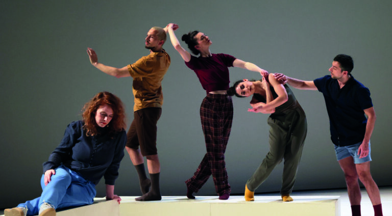 Vijf dansers zijn in verschillende poses te zien op een minimalistisch podium met een neutrale achtergrond.