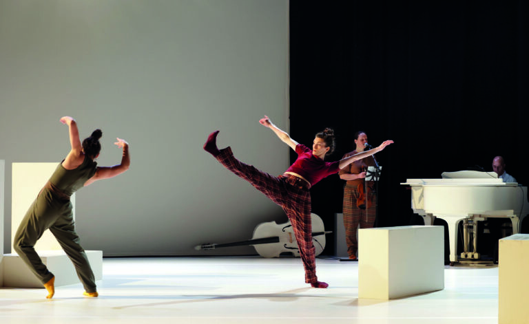 Drie dansers voeren een choreografie uit op een minimalistisch podium met een blanco achtergrond.