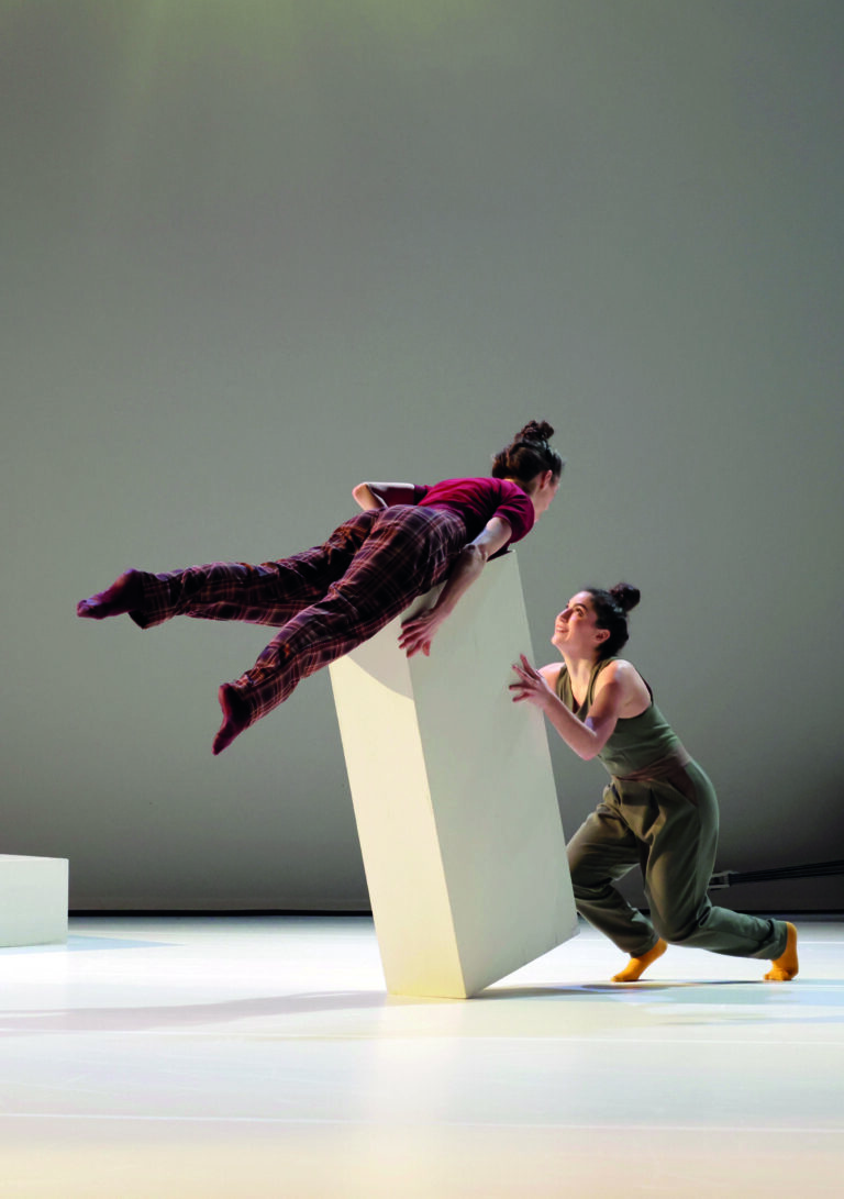 Twee dansers voeren een fysieke act uit waarbij één danser in de lucht zweeft boven een verticale witte rechthoek.