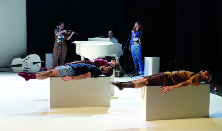 Een artistieke uitvoering met dansers die op zwarte kisten liggen, terwijl een muzikant op een piano speelt en een andere muzikant viool speelt.