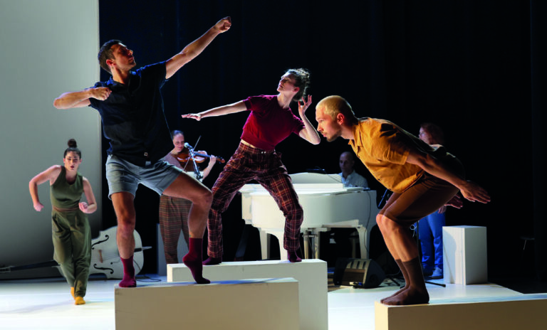 Twee dansers en een muzikant voeren een levendige performance uit op verhoogde platforms met een piano op de achtergrond.