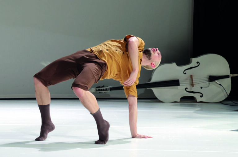 Een danser voert een acrobatische beweging uit op een witte vloer, met een grote strijkinstrument op de achtergrond.