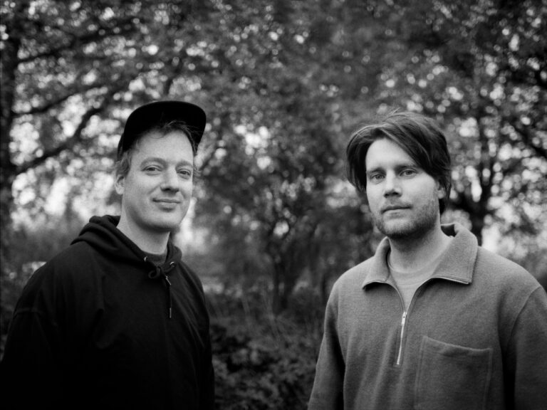 Twee mannen staan samen in een groene omgeving, met bomen op de achtergrond.