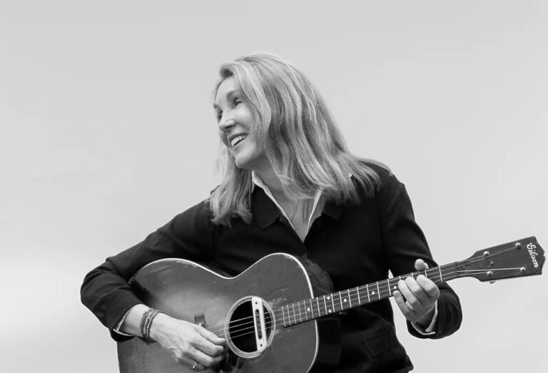 Zwart wit foto van zangeres Diana Jones, ze zit voor een witte muur en kijkt naar links met een acoustische gitaar in haar hand.