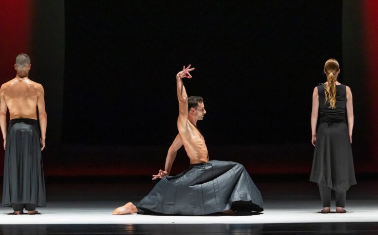 Een danser in een zwart kostuum voert een dynamische beweging uit op een podium met een donkere achtergrond.