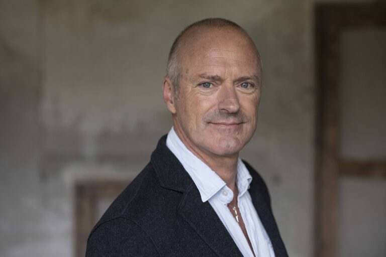 Een man met kort, grijs haar, gekleed in een zwart jasje en een witte shirt, staat glimlachend voor een neutrale achtergrond.