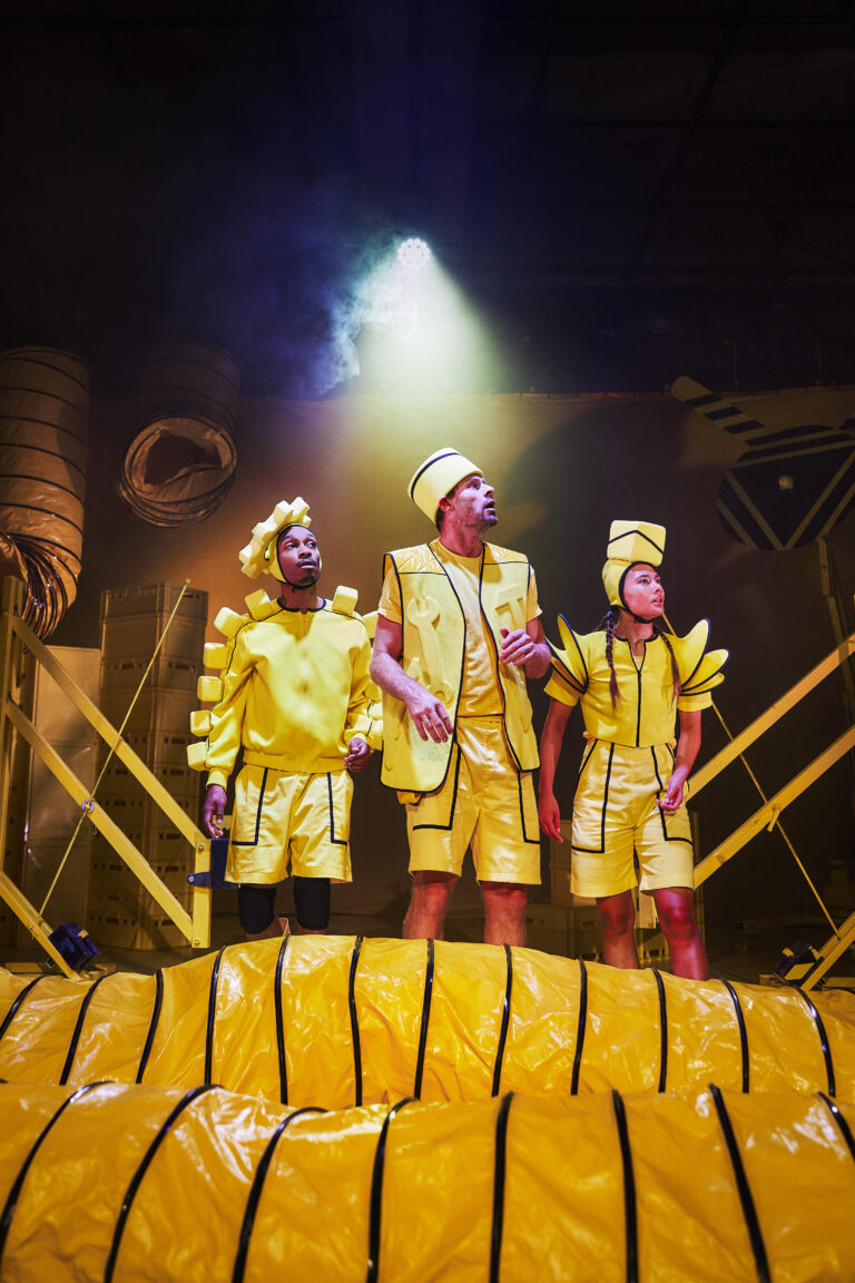 Drie performers in opvallende gele kostuums staan op een podium, omgeven door theatrale lichteffecten en decor.