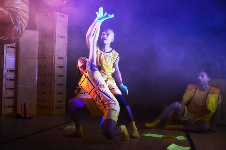 Twee dansers in gele kostuums bewegen onder kleurrijke lichteffecten en mist.