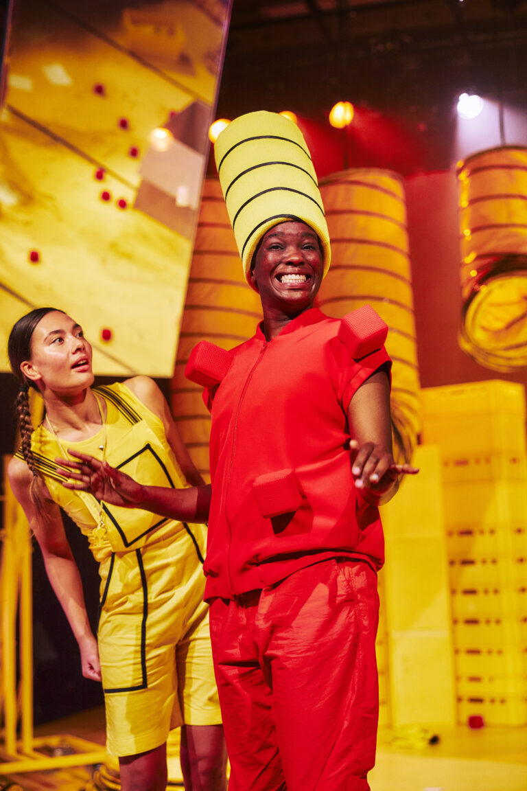 Twee performers op een kleurrijk podium, één in een opvallende gele hoed en rode outfit, en de ander in een gele jurk.