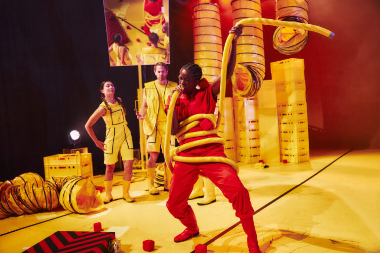 Drie performers in een kleurrijke, theatrale setting, waarbij één performer bezig is met het balanceren van gele slangen en de andere twee toekijken.