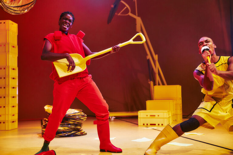 Performance waarbij een artiest in een felrode outfit een gele schep als gitaar bespeelt, met op de achtergrond verschillende obiecten.