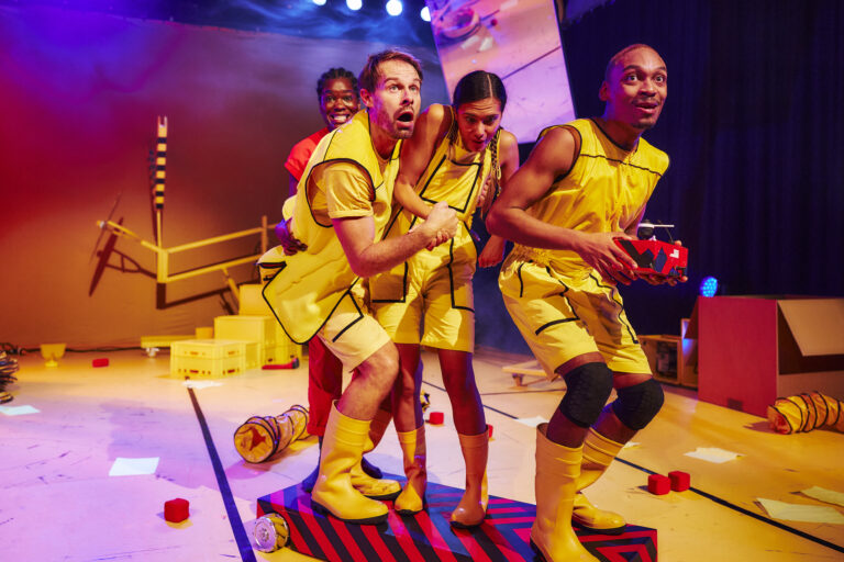 Vier acteurs in gele kostuums poseren samen op een speelse theaterdecor met kleurrijke elementen.