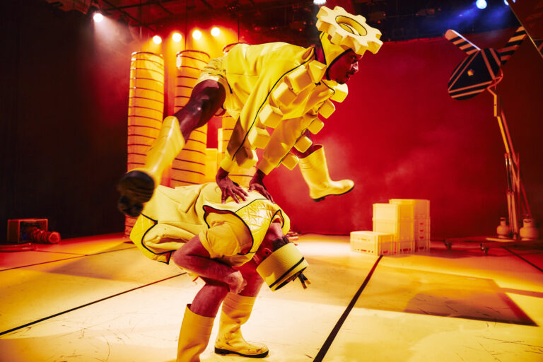 Een acrobat in een geel kostuum voert een spectaculaire stunt uit op een podium.