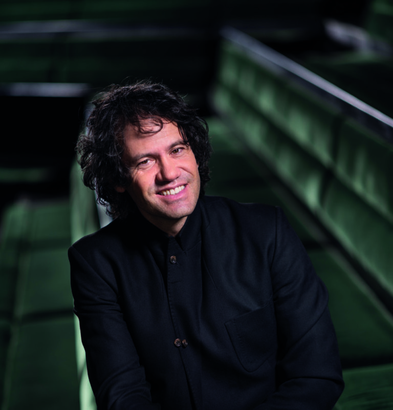 Een man met krullend haar en een zwarte jas zit in een zaal met groene stoelen en glimlacht.