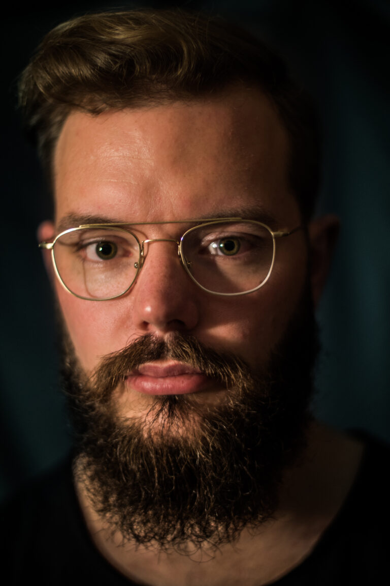 Portret van een man met een volle baard en een bril, gefotografeerd met een donkere achtergrond.