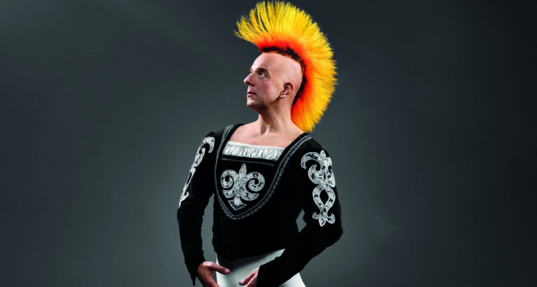 Een persoon met een opvallende gele en oranje mohawk en een zwart kledingstuk met zilveren versieringen poseert artistiek.